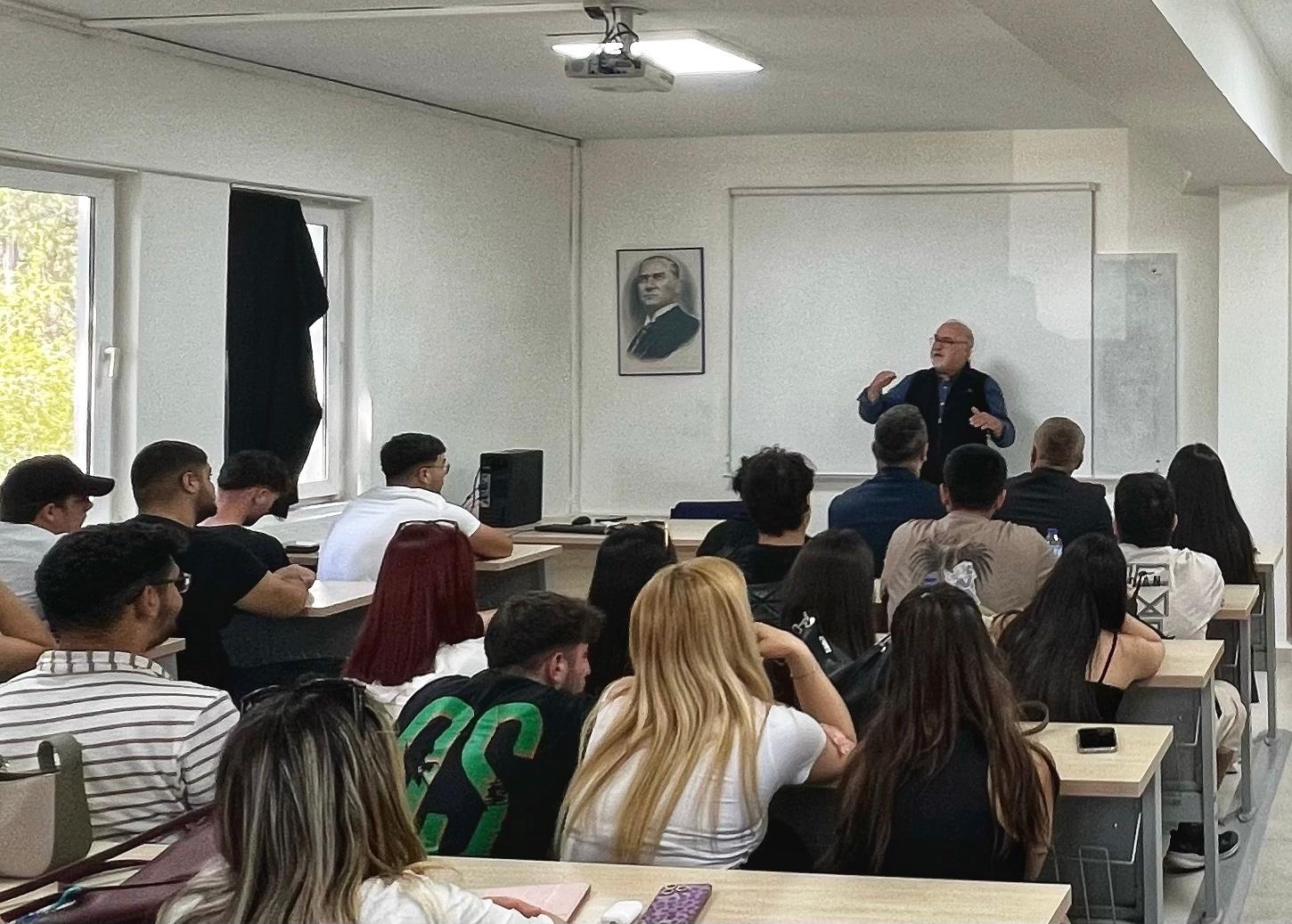 Bir sınıf ortamında, öğrencilere ders anlatan orta yaşlı bir erkek öğretmen ayakta konuşma yapıyor. Öğrenciler sıralarda oturmuş, dikkatle öğretmeni dinliyor. Sınıfın ön duvarında bir beyaz tahta, üstünde Atatürk portresi ve tavanda bir projeksiyon cihazı bulunuyor. Sınıf aydınlık ve modern bir yapıya sahip.